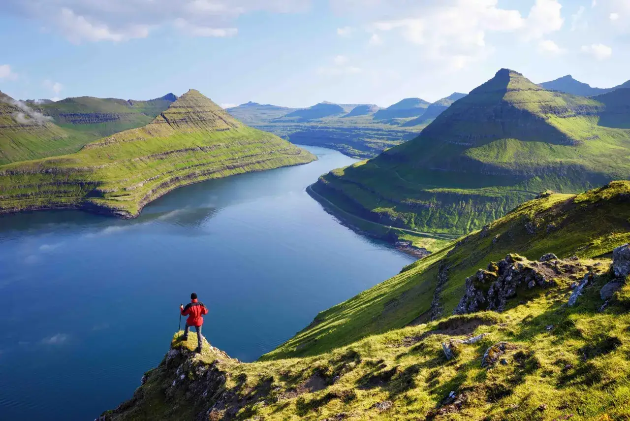 Un randonneur admire un paysage spectaculaire des îles Féroé, avec des montagnes verdoyantes et un fjord bleu.