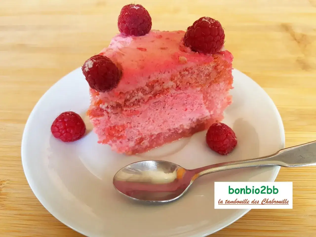 Une part de charlotte aux framboises, garnie de fruits rouges frais, sur une assiette blanche avec une cuill&egrave;re.