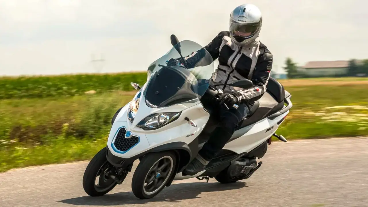 Biały Piaggio MP3 500 z kierowcą w kasku i kurtce jedzie po drodze. Nawet z jego zaletami, piaggio mp3 500 wady mogą być istotne.
