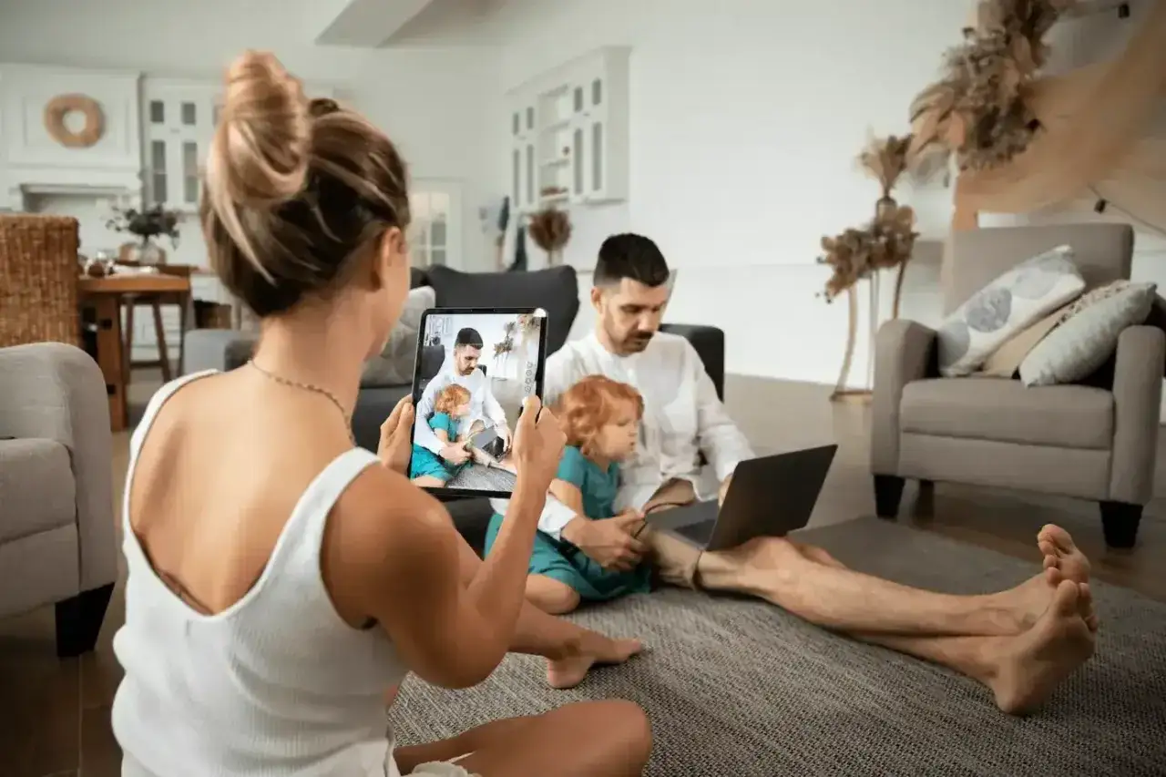 Familia interactuando con pantallas. Un padre trabaja en su portátil mientras la madre graba un vídeo de su hijo y el padre en la tablet.
