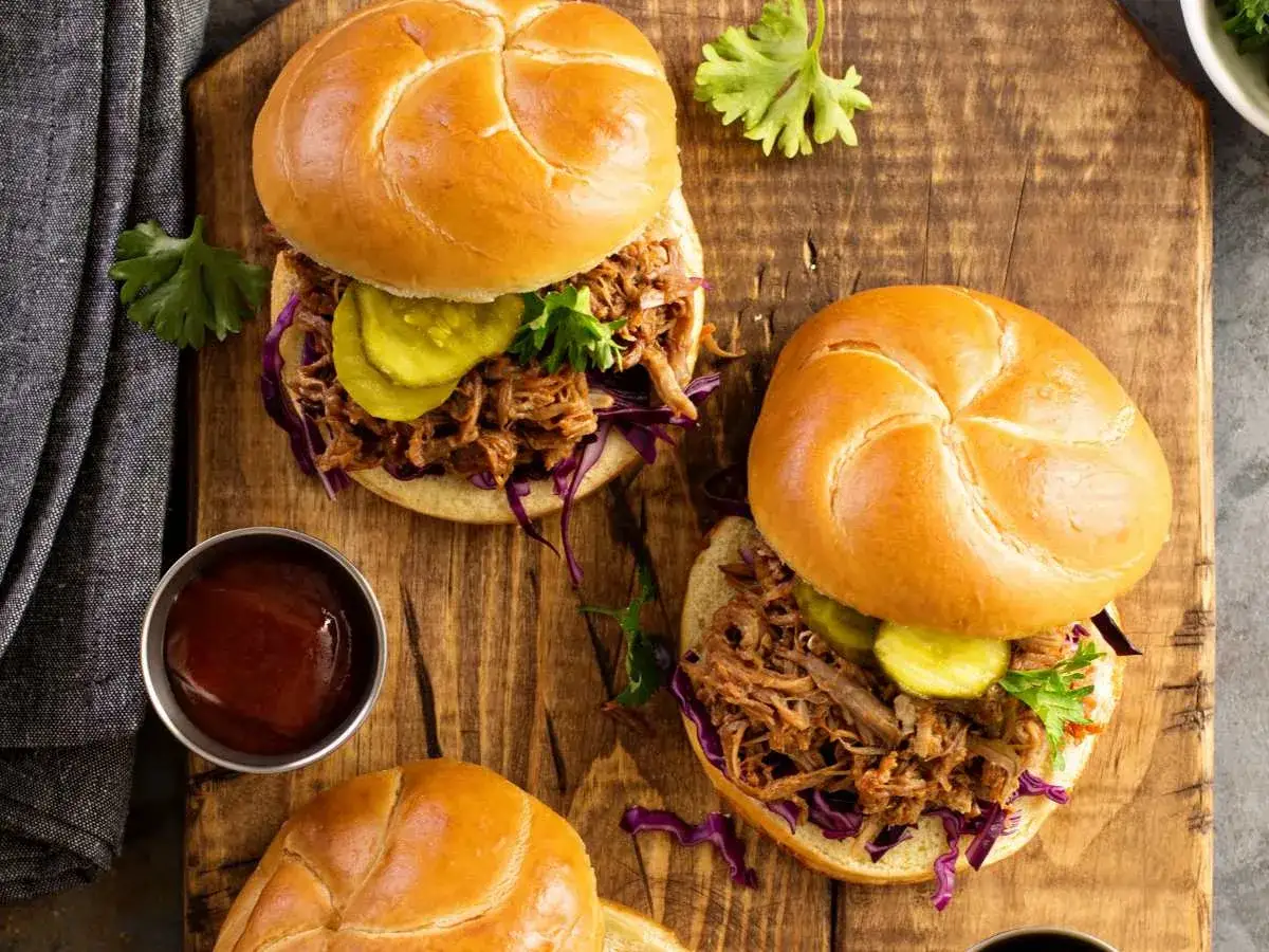 Dos deliciosos s&aacute;ndwiches de pulled pork con repollo morado y pepinillos, servidos en una tabla de madera r&uacute;stica.