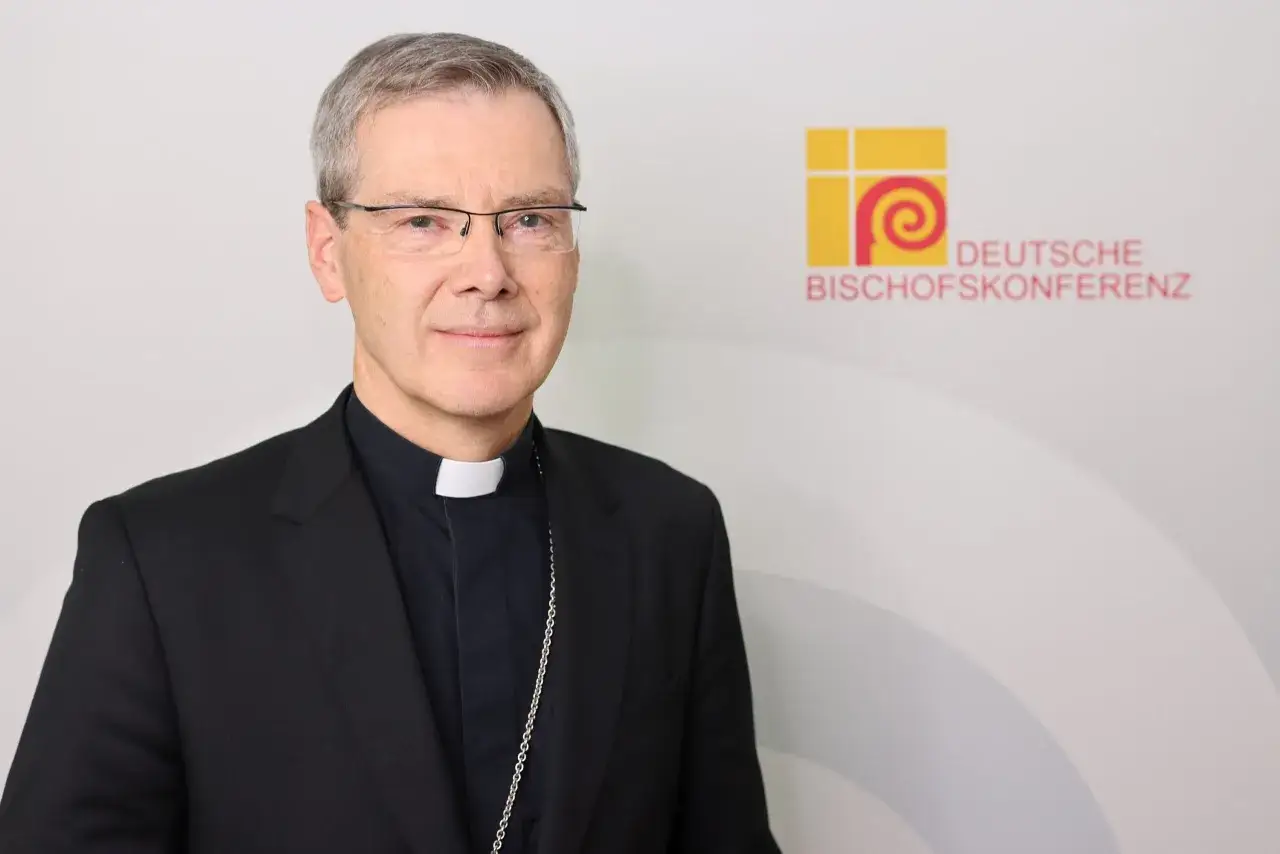 Ein Mann mit Brille und geistlichem Beffchen, im schwarzen Talar, steht vor dem Logo der Deutschen Bischofskonferenz.