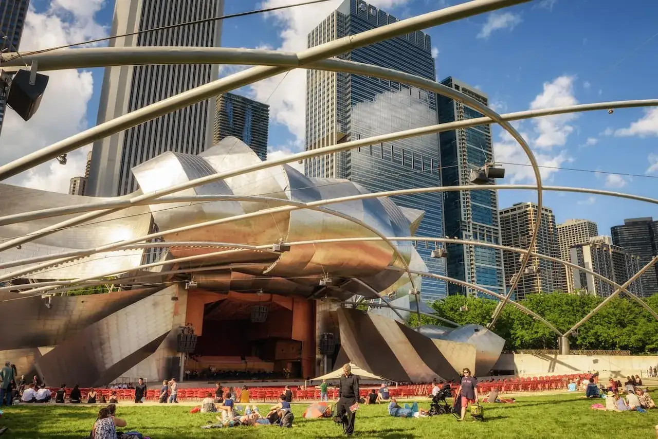 Ludzie odpoczywają na trawie przed sceną w Millennium Park, otoczeni nowoczesną architekturą i drapaczami chmur.
