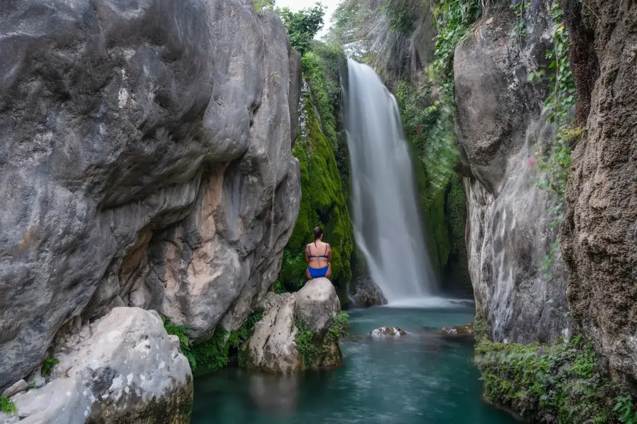 Una mujer sentada en una roca contempla la cascada de las fuentes del Algar, rodeada de vegetación y rocas.