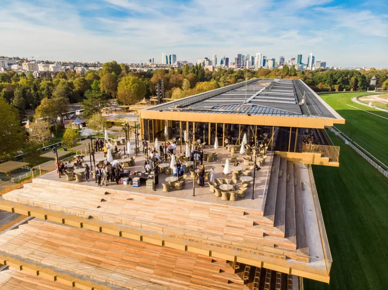Vue aérienne du toit d'un bâtiment moderne avec des tables et des parasols sur une terrasse, avec le paysage urbain de Paris et l'hippodrome de Longchamp en arrière-plan.