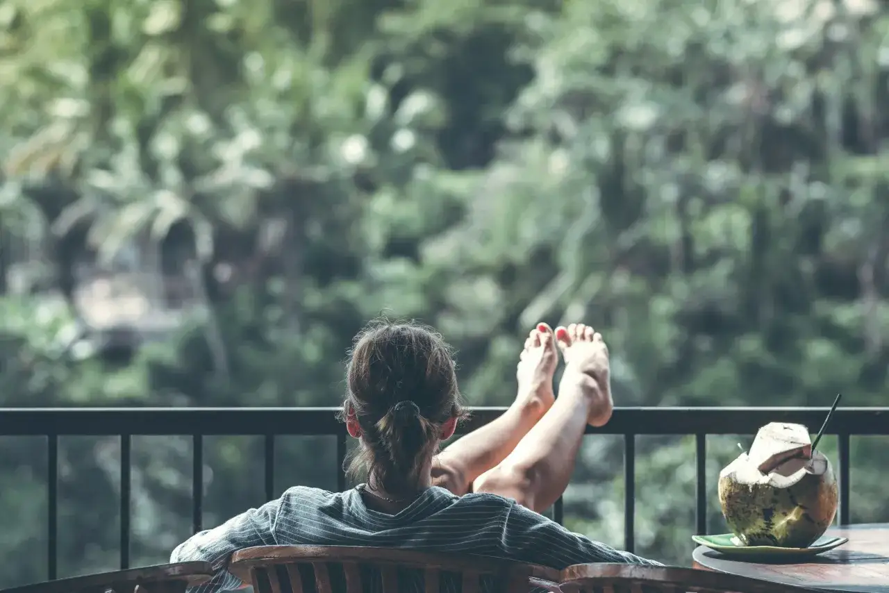 A woman with her feet up on a balcony railing, a coconut drink nearby, enjoying a moment to decompress amidst lush greenery.