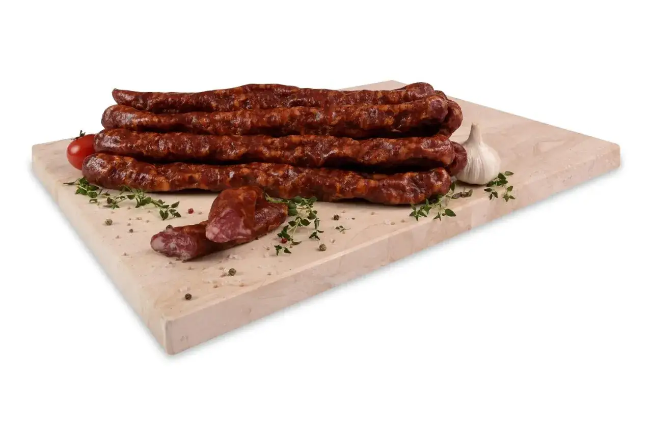Kilka długich, wędzonych kiełbasek, z których jest salami, ułożonych na desce z czosnkiem i pomidorkiem.