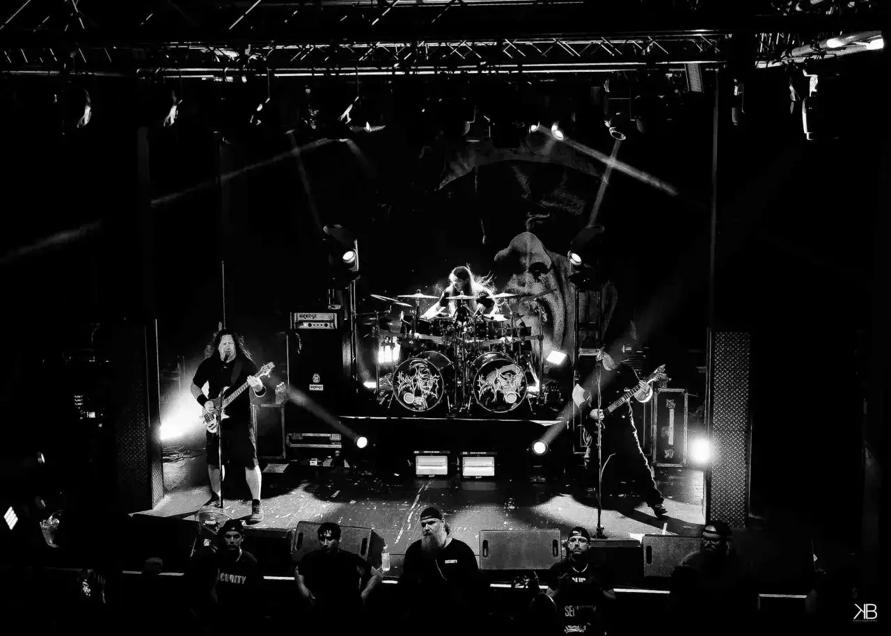 Groupe de metal sur sc&egrave;ne, &eacute;clairage dramatique. Le batteur est au centre, entour&eacute; de symboles sombres rappelant un f&oelig;tus mourant.