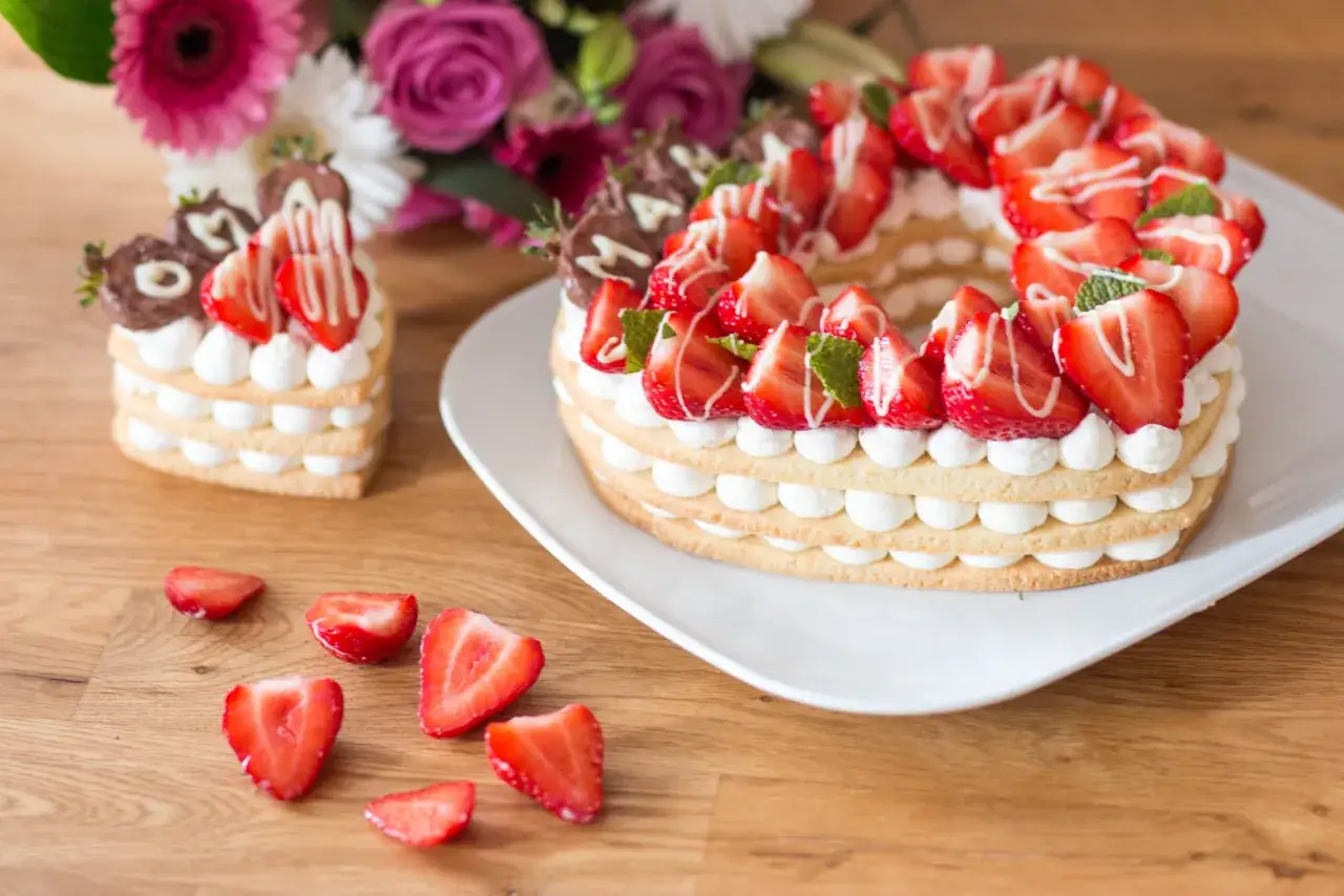 Zwei kunstvolle Kuchen mit viel Creme und Erdbeeren, ein kleiner Herz-Kuchen und ein gro&szlig;er runder Kuchen, daneben frische Erdbeerst&uuml;cke.