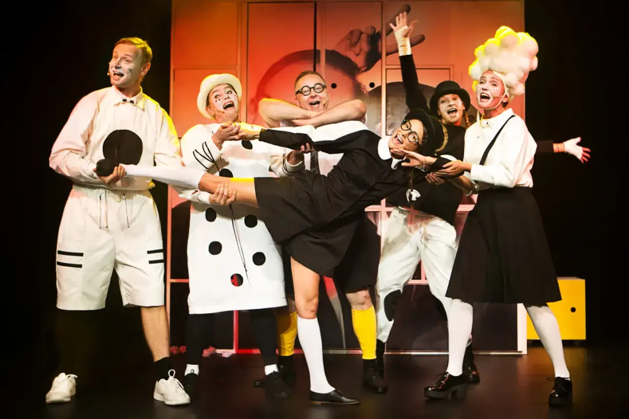 Wesoły spektakl teatralny dla dzieci. Aktorzy w kostiumach w grochy i w białych strojach bawią się na scenie, tworząc radosną atmosferę.