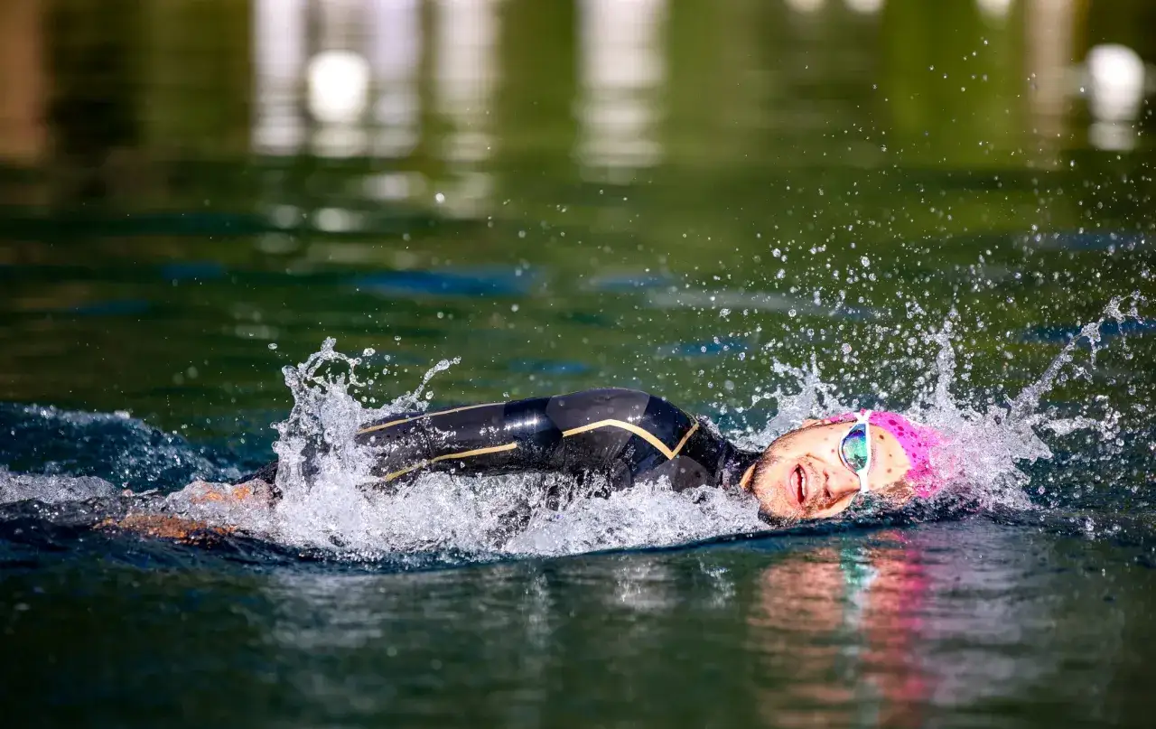En tävlingssimmare i en våtdräkt kämpar sig framåt i vattnet, med ansiktet vänt mot kameran. Vattenstänk och ansträngning syns tydligt under denna ironman simning.