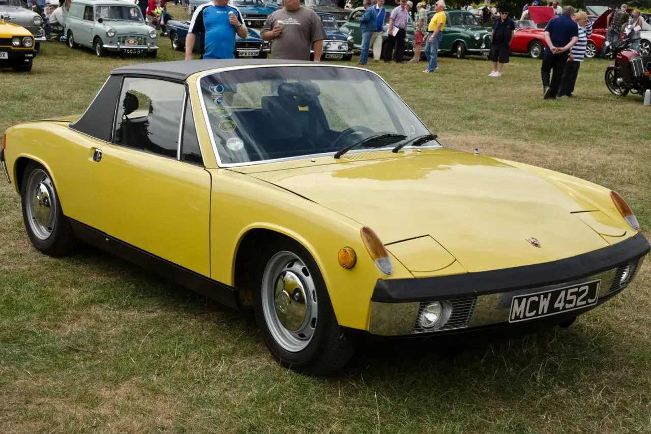 Żółty Porsche 914 z czarnym dachem, zaparkowany na trawie wśród innych klasycznych samochodów i ludzi.