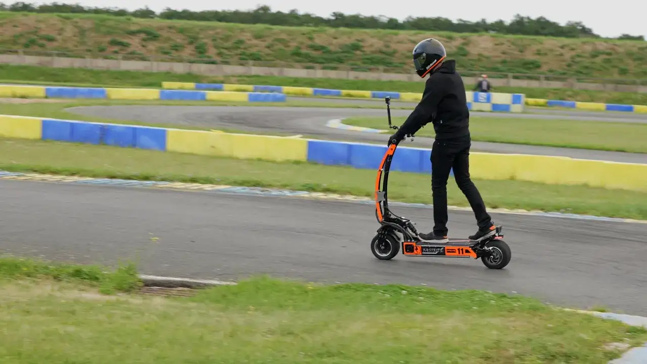Un homme en casque et tenue noire roule sur une trottinette électrique orange sur un circuit. Il semble vouloir debrider trottinette pour plus de vitesse.