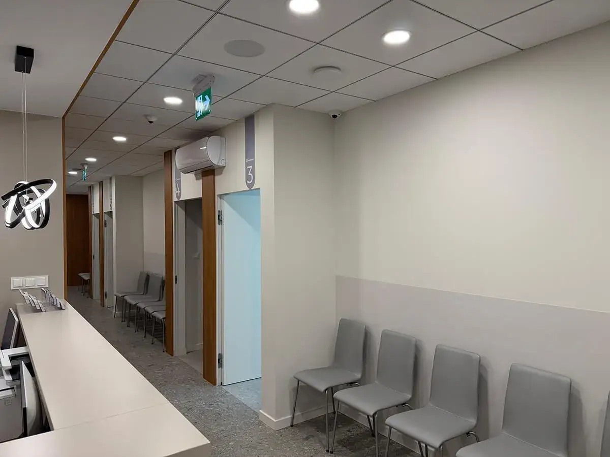 Nowoczesna poczekalnia kliniki z ladą recepcyjną, rzędami krzeseł i drzwiami gabinetów.