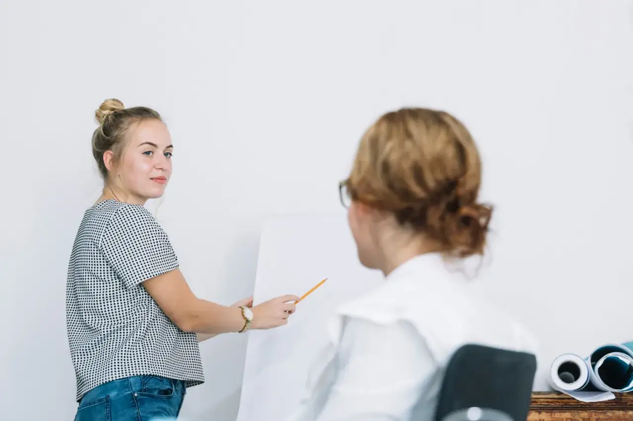 Młoda kobieta z blond włosami upiętymi w kok, w bluzce w kropki, wskazuje ołówkiem na białą tablicę. Sesja psychoterapii poznawczo-behawioralnej.