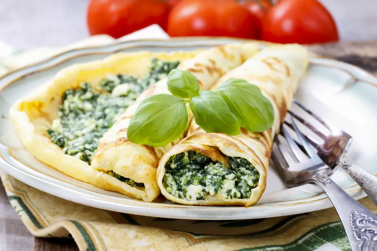 Naleśniki ze szpinakiem i serem feta na obiad, udekorowane listkami bazylii. Obok widoczne widelce i pomidory.