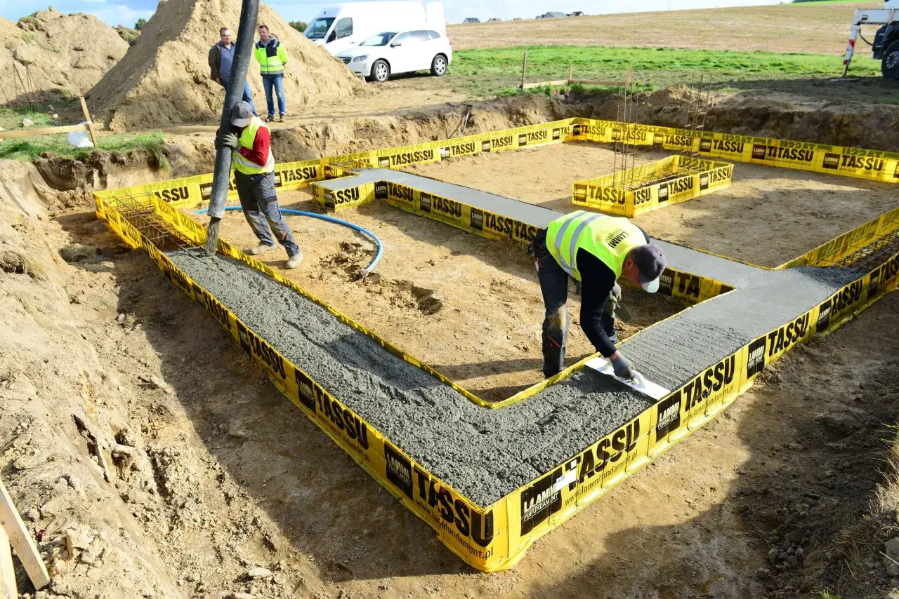 Betonowanie ław fundamentowych. Robotnicy wylewają beton, a jeden z nich wygładza powierzchnię. Cena robocizny jest kluczowa dla budowy.