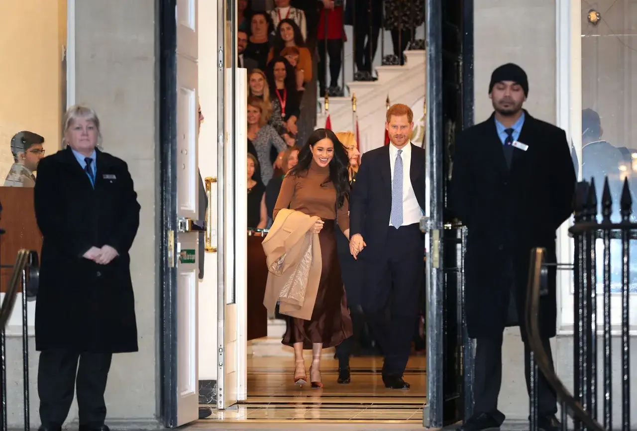 Meghan och Harry går ut ur en byggnad, omgivna av säkerhetspersonal och en folkmassa.