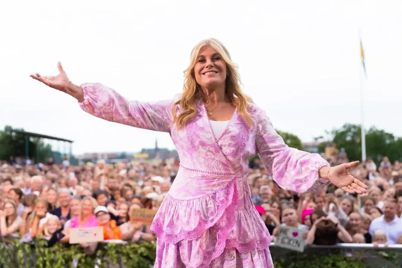 En glad kvinna i rosa klänning med utsträckta armar, publik i bakgrunden. Allsång på Skansen i full gång.