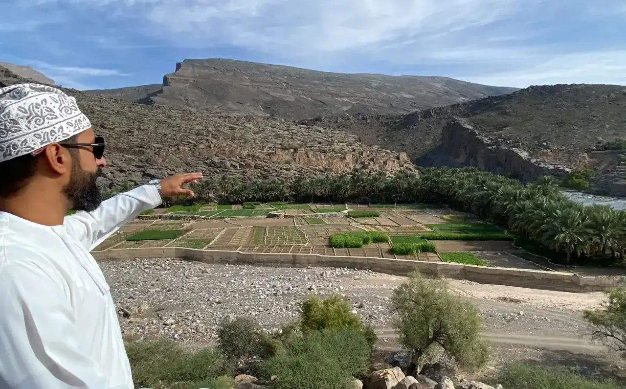 Mężczyzna w tradycyjnym stroju wskazuje na zielone pola uprawne w dolinie. Czy Oman jest bezpieczny? Tak, to spokojny kraj z piękną przyrodą.