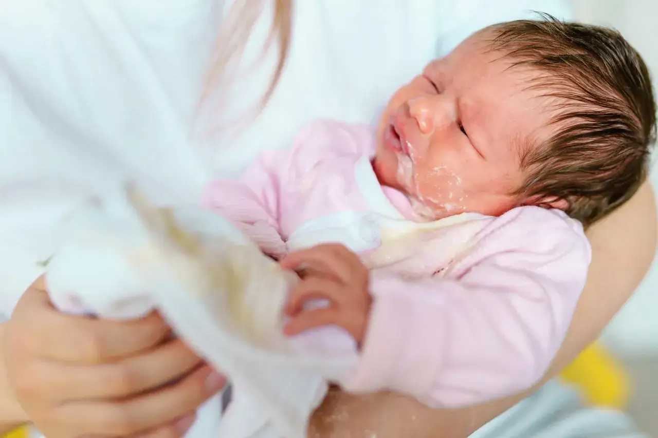 Malutkie dziecko w r&oacute;żowym ubranku, z widocznym śladem po jedzeniu na buzi, co może sugerować refluks u niemowlaka. Mama wyciera mu buzię.