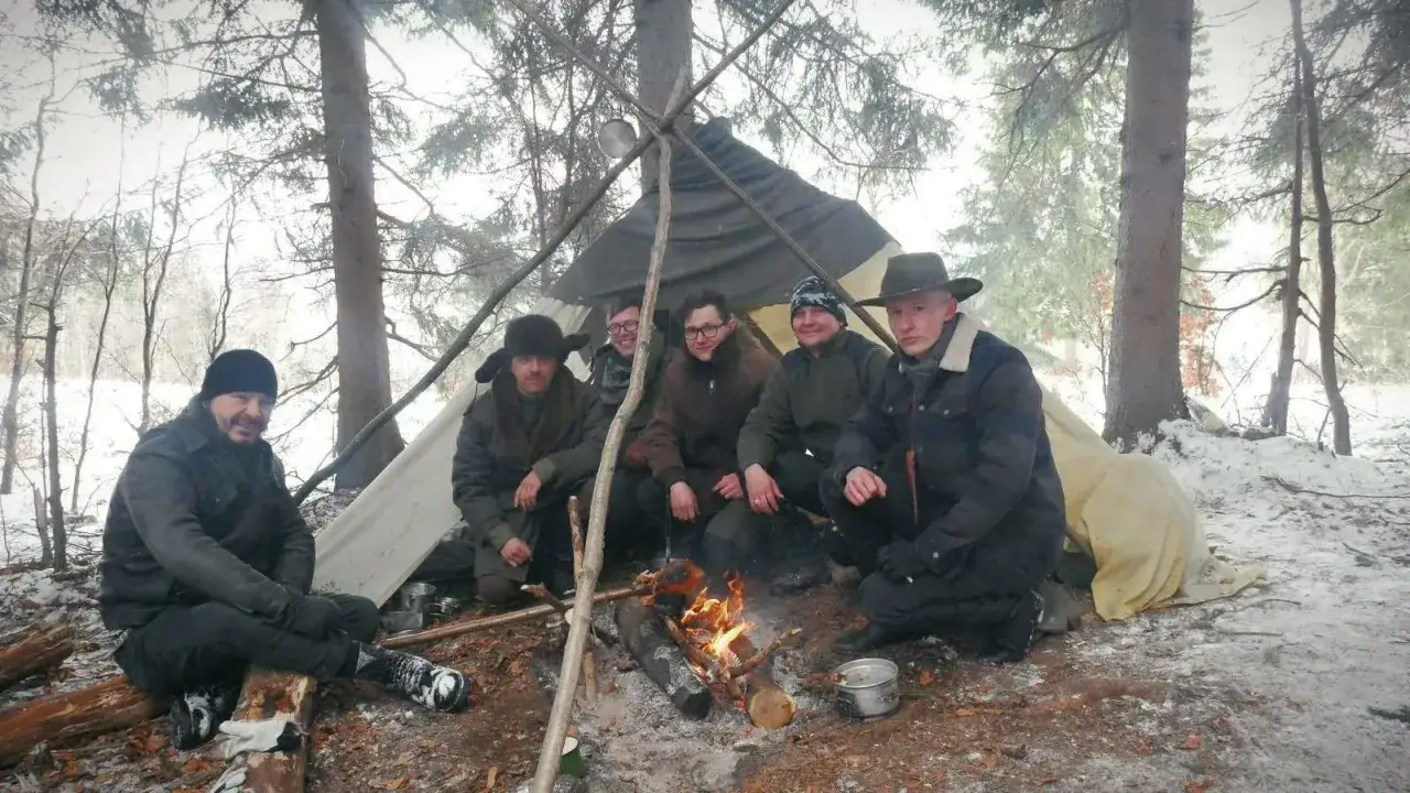 Grupa mężczyzn siedzi przy ognisku pod namiotem w lesie. To jest prawdziwy **outdoor** – przygoda i bliskość natury.