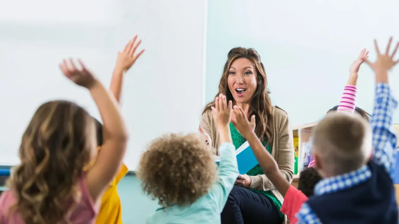 Maestra animada interactúa con niños que levantan las manos, participando en divertidos juegos de lengua en el aula.