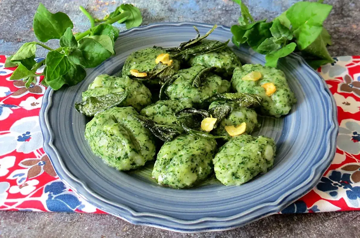 Zielone gnocchi ze szpinakiem, polane masłem z szałwią i czosnkiem, podane na niebieskim talerzu.