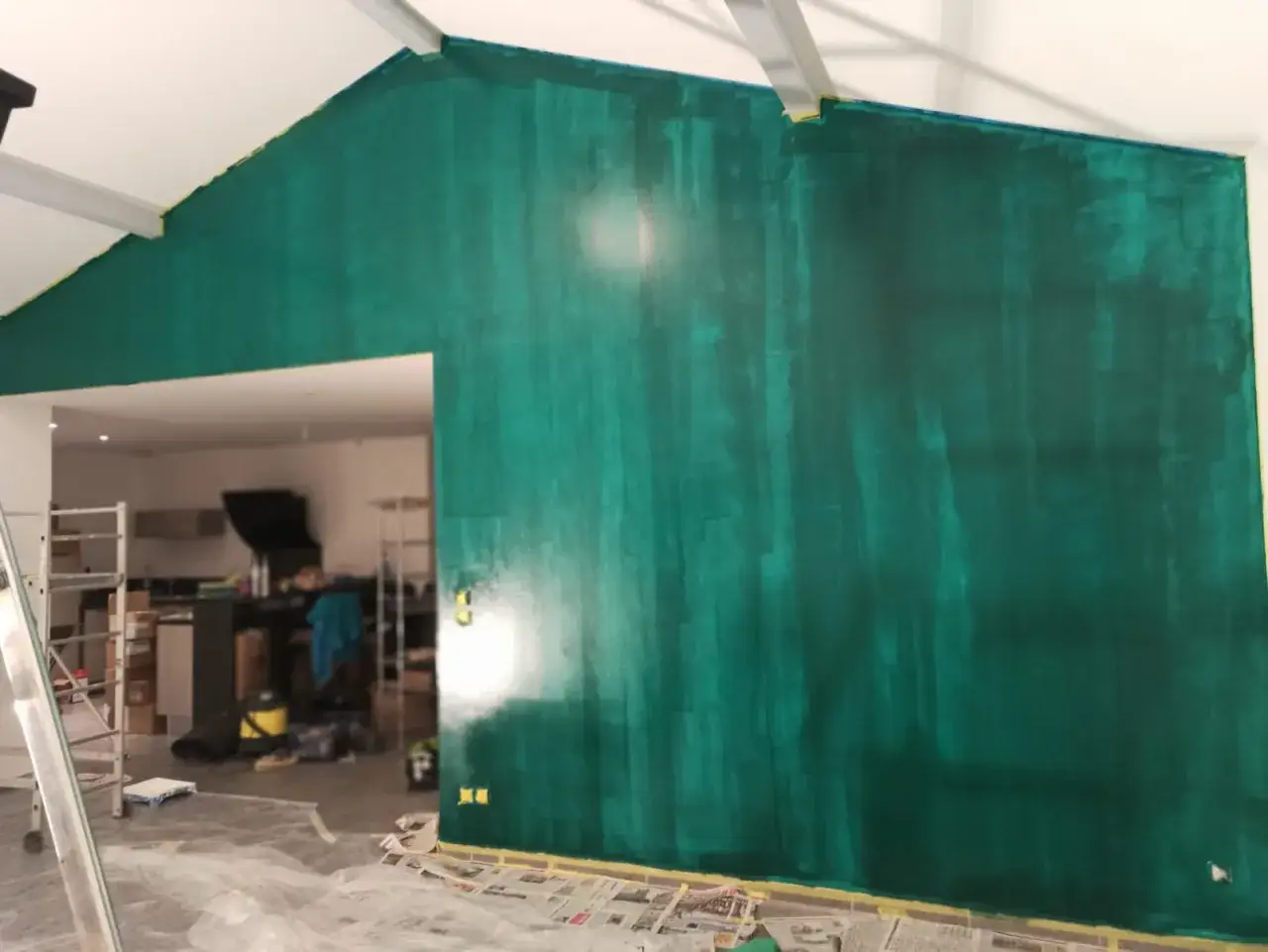 Mur vert profond, fraîchement peint avec 2 ou 3 couches pour un effet texturé. Le chantier est en cours, avec du matériel de peinture au sol.