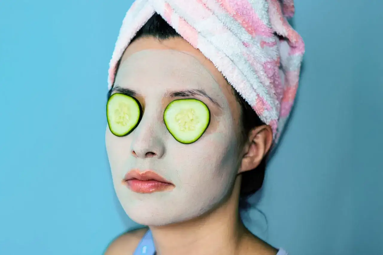 Mujer con mascarilla facial y rodajas de pepino en los ojos, envuelta en una toalla rosa y blanca.