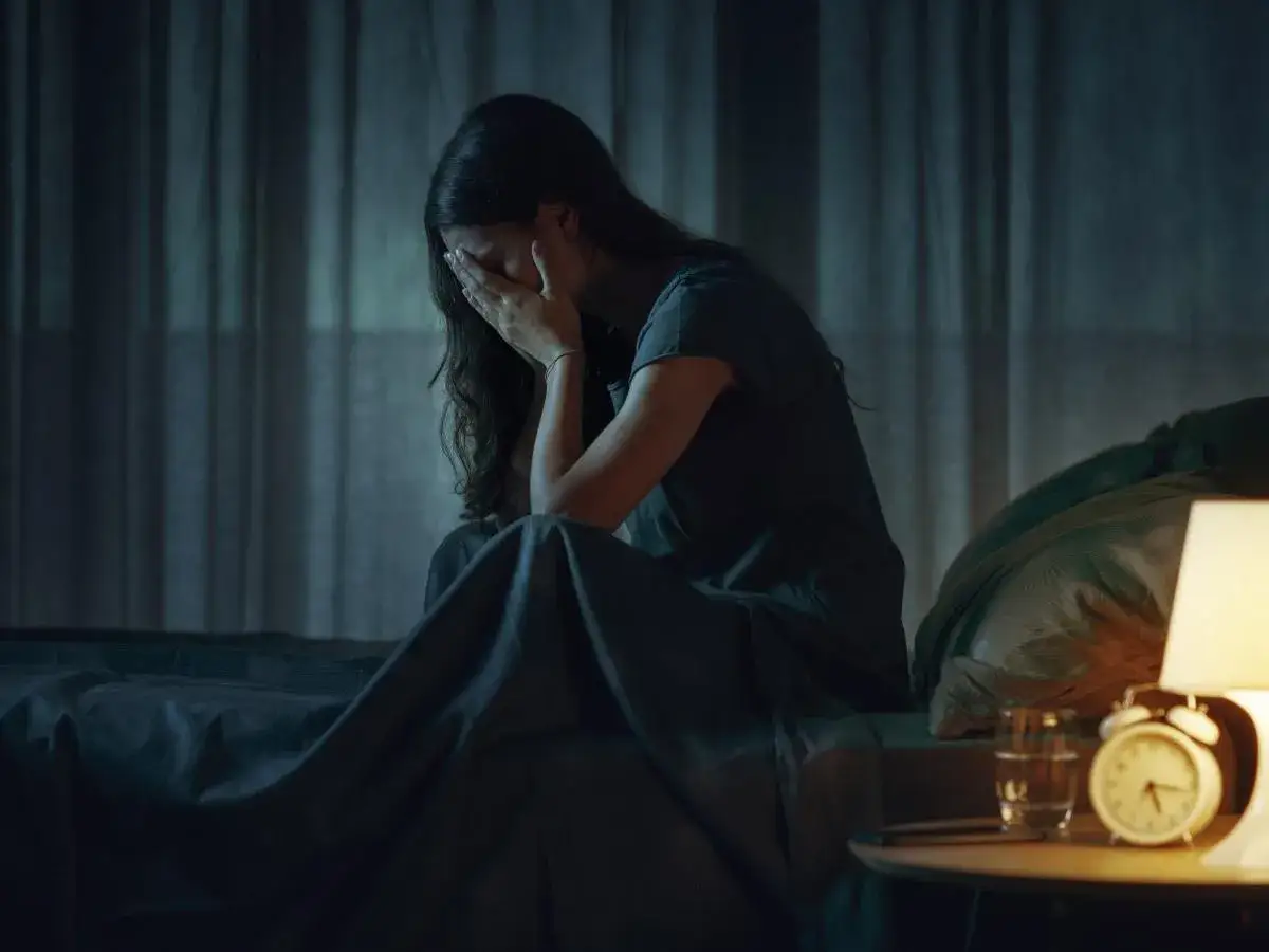 Mujer sentada en la cama, con la cara entre las manos, sufriendo la vigilia del sueño. La noche es oscura, solo iluminada por una lámpara.