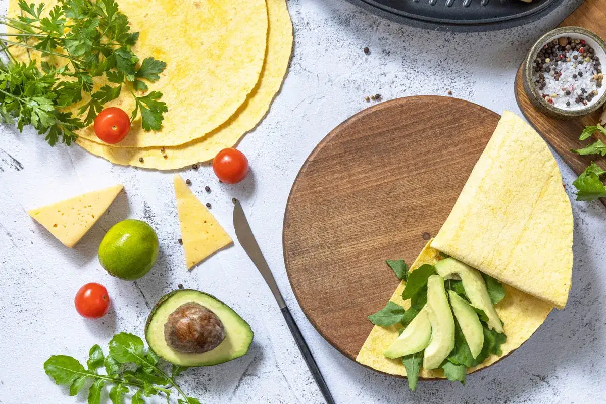 Keto tortilla z siemienia lnianego, przygotowana z awokado i rukolą, obok pomidorków, sera i limonki.