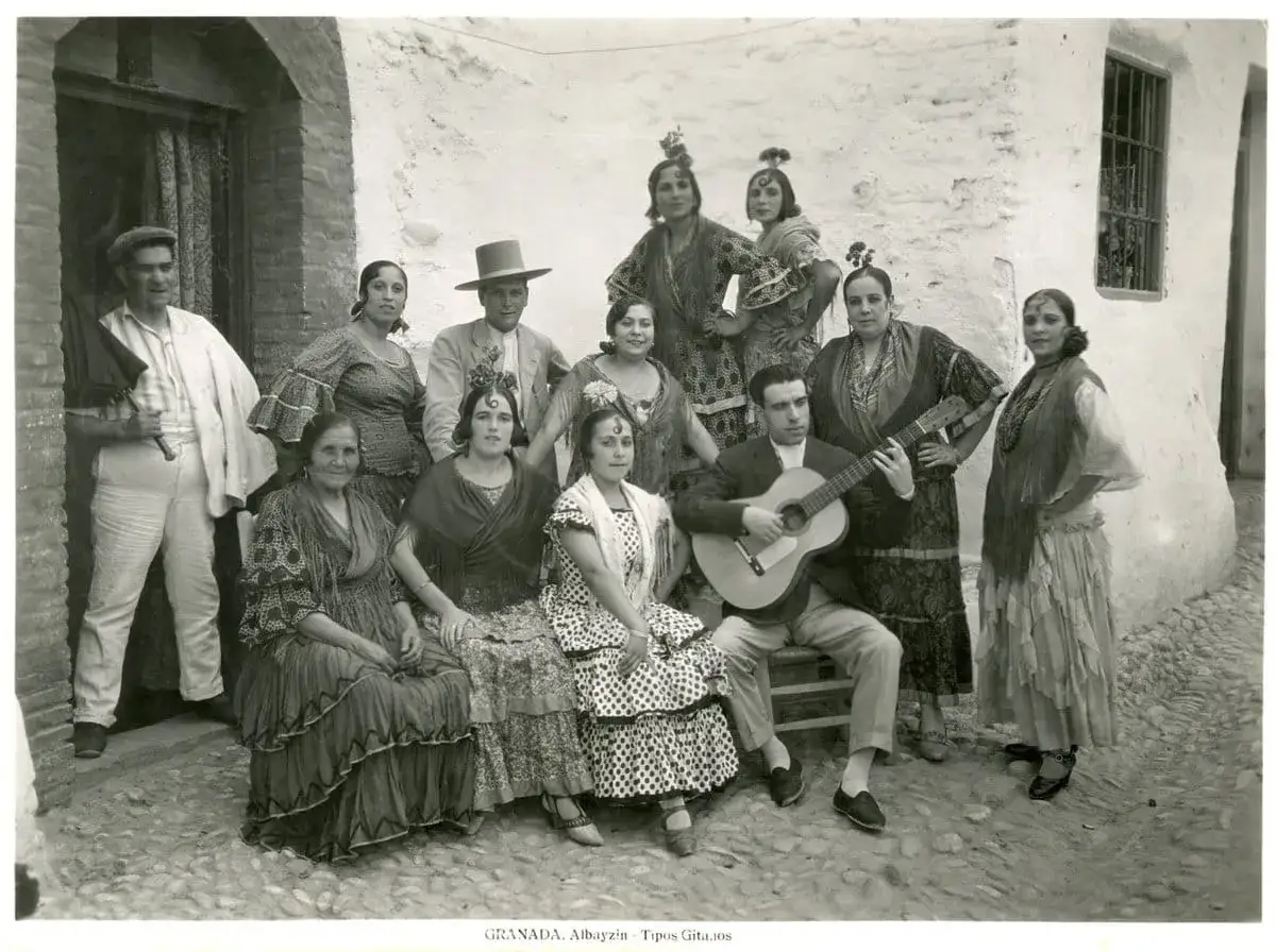 Grupo de personas vestidas con trajes flamencos, algunos con mantones y peinetas. Un hombre toca la guitarra. Los nombres flamencos de estas figuras resuenan en la tradición.