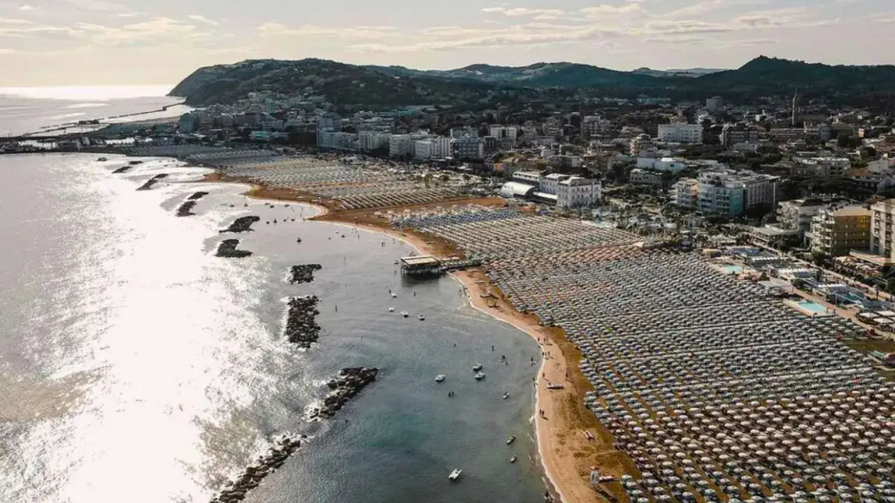 Vista aerea del porto di Cattolica con la spiaggia affollata di ombrelloni e lettini, mare calmo e colline sullo sfondo.