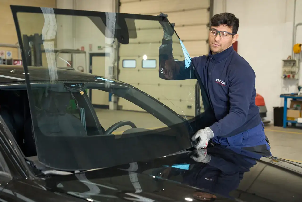 Tecnico con occhiali protettivi rimuove un parabrezza rotto da un'auto nera in officina.