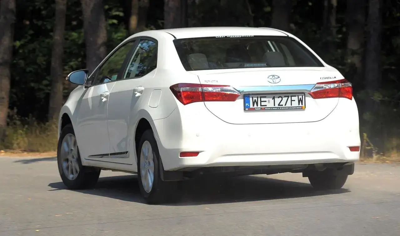 Biała Toyota Corolla E16 z tablicą WE 127FW jedzie drogą wśród drzew.