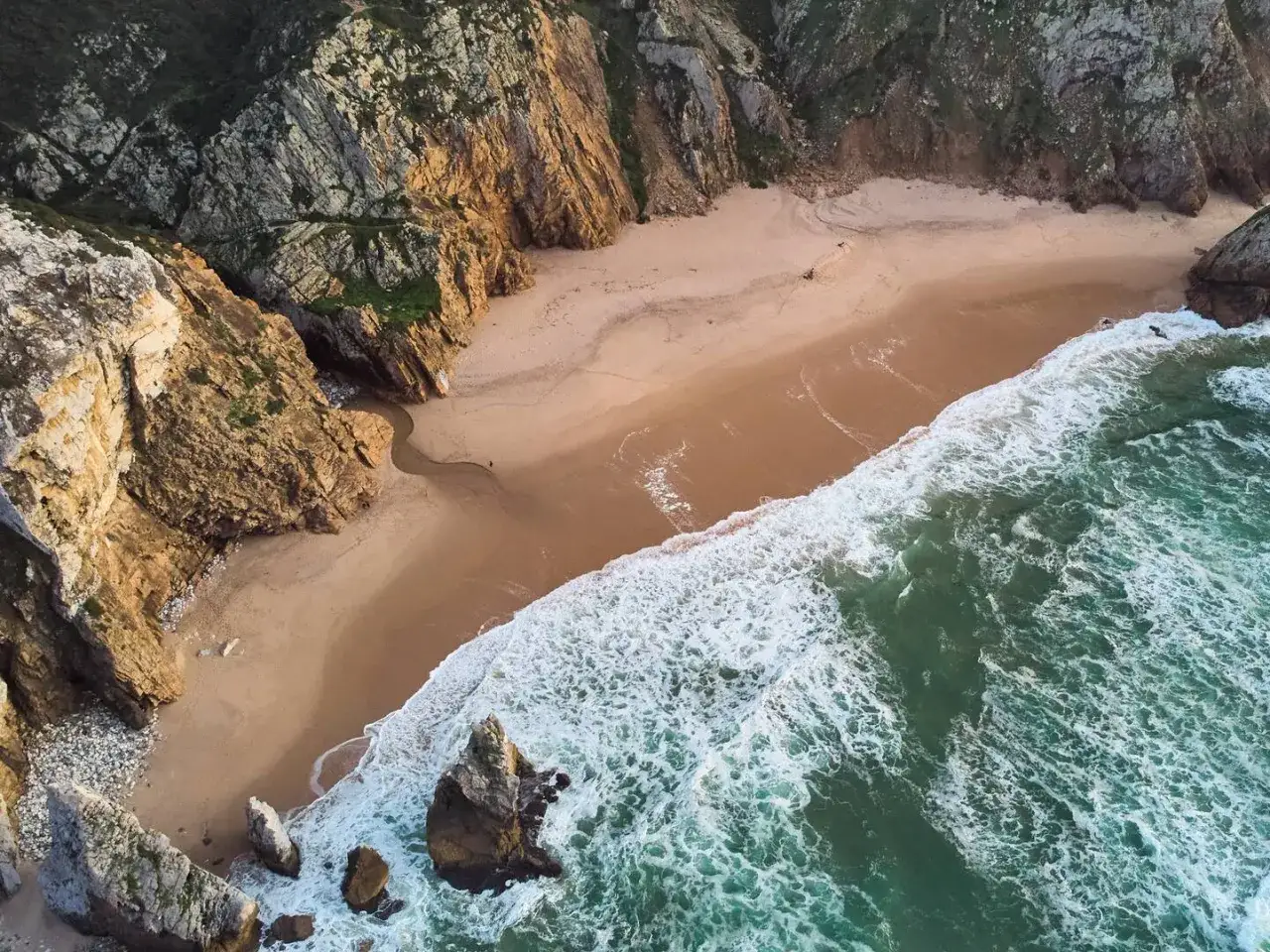Atemberaubende **Lissabon Natur**: Eine einsame Bucht mit goldenem Sand, umgeben von steilen Felsen und tosenden Wellen. Ein kleiner Bach schl&auml;ngelt sich zum Meer.