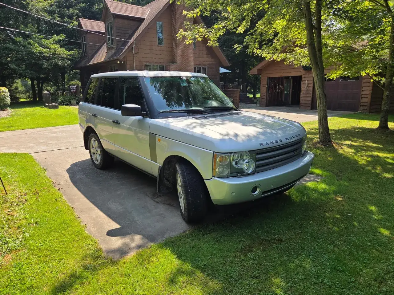 Silberner Range Rover Sport parkt vor einem Holzhaus. M&ouml;glicherweise gibt es Probleme mit der Luftfederung, da das Fahrzeug etwas tief steht.