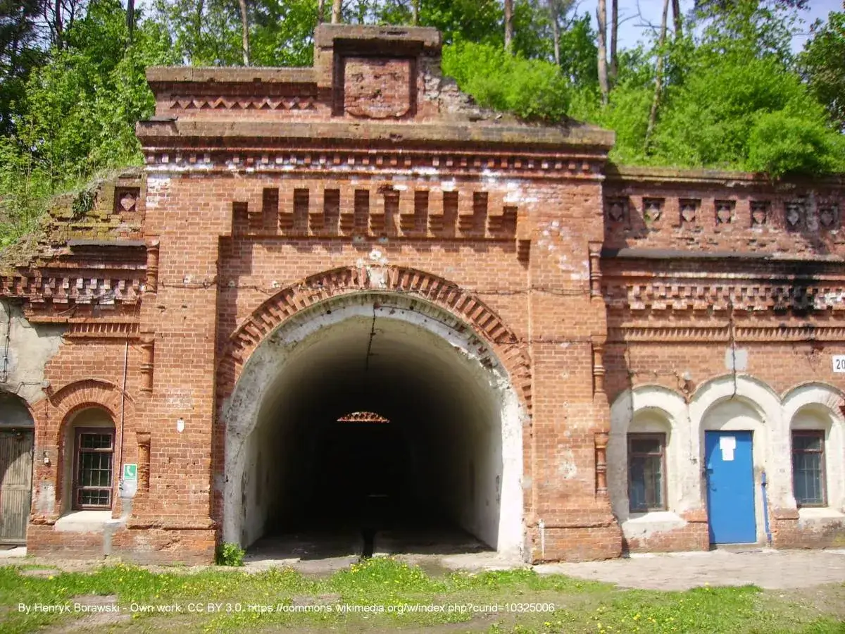 Fortyfikacje w Osowcu: masywny ceglany łuk wejściowy do tunelu, z małymi okienkami i niebieskimi drzwiami po bokach.