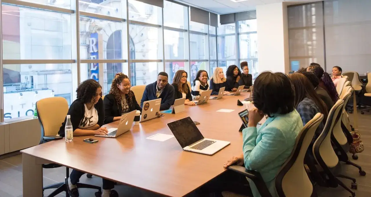 A diverse group of professionals in a modern boardroom, discussing how to get board positions. Laptops and tablets are on the table.