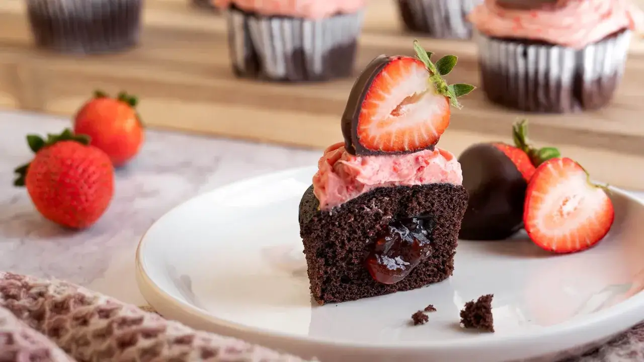 En skuren choklad cupcake med rosa frosting, fylld med sylt och toppad med en chokladdippad jordgubbe. Bredvid ligger fler choklad cupcakes och f&auml;rska jordgubbar.