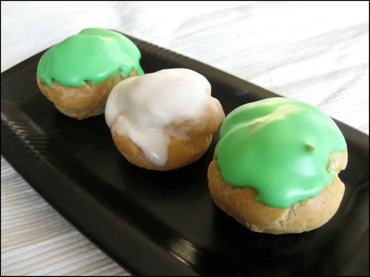 Trois choux à la crème, deux verts et un blanc, disposés sur une assiette noire.