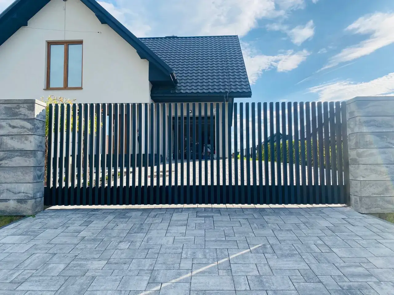 Nowoczesna, czarna brama przesuwna z pionowymi lamelami, idealnie komponuje się z kamiennymi słupkami i podjazdem z kostki. Zastanawiasz się, ile kosztuje brama przesuwna z montażem?