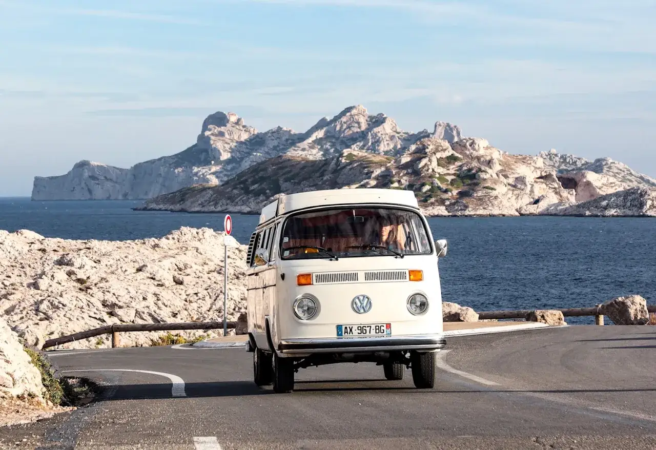 Un camping-car Volkswagen blanc roule sur une route c&ocirc;ti&egrave;re avec des falaises rocheuses et la mer en arri&egrave;re-plan, une belle journ&eacute;e pour explorer Marseille en camping-car.