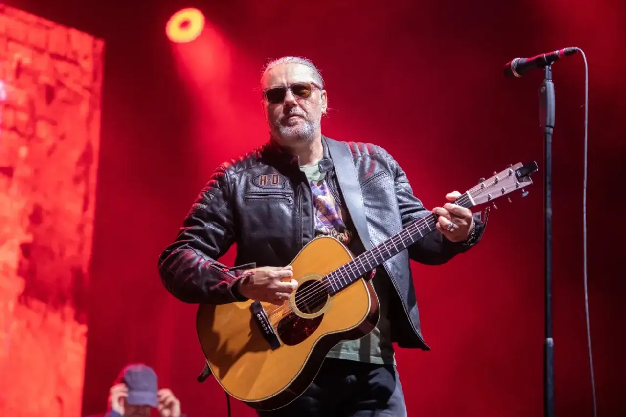 Dżem wokalista z gitarą na scenie, w sk&oacute;rzanej kurtce i okularach przeciwsłonecznych.