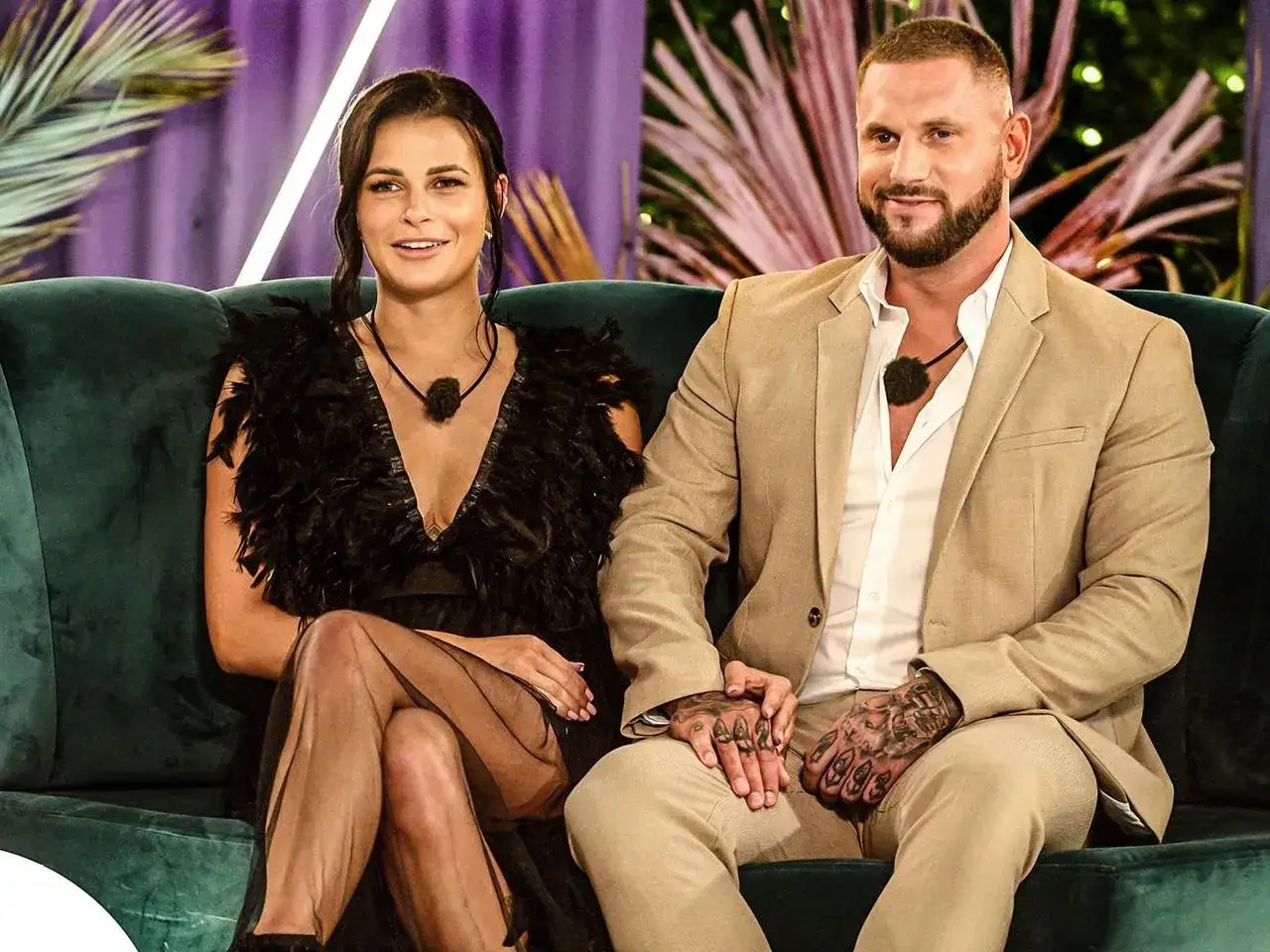Sara z Love Island w czarnej, pióropuszowej sukni i jej partner siedzą na kanapie.