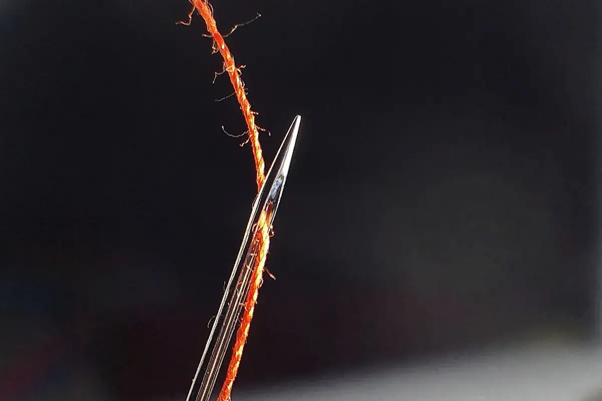 Gros plan d'une aiguille microtex traversant un fil orange. Idéale pour les tissus délicats.