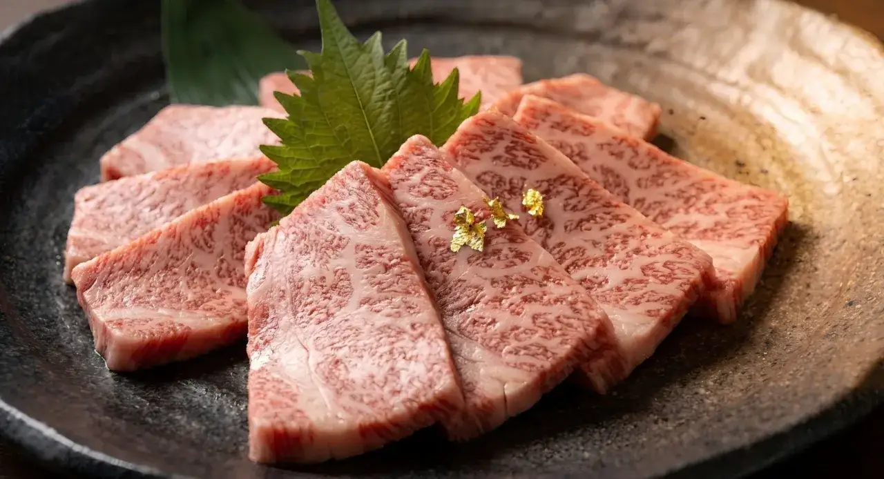 Filetes de carne japonesa cara, con marmoleado intenso y un toque de pan de oro, servidos en un plato oscuro con hojas verdes.