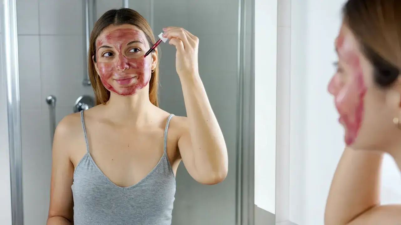 Kobieta z czerwoną maską na twarzy aplikuje kolejne krople serum. To jej rytuał z kwasami na twarz dla promiennej cery.
