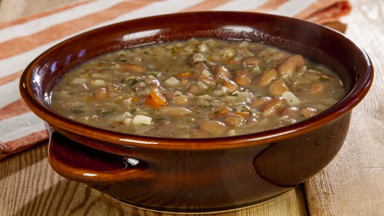 Una ciotola di terracotta marrone piena di saporita minestra di fagioli, con pezzi di verdure e erbe aromatiche.